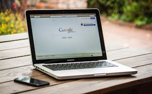 A MacBook Pro displaying Google Search on a wooden table outdoors, next to a smartphone.